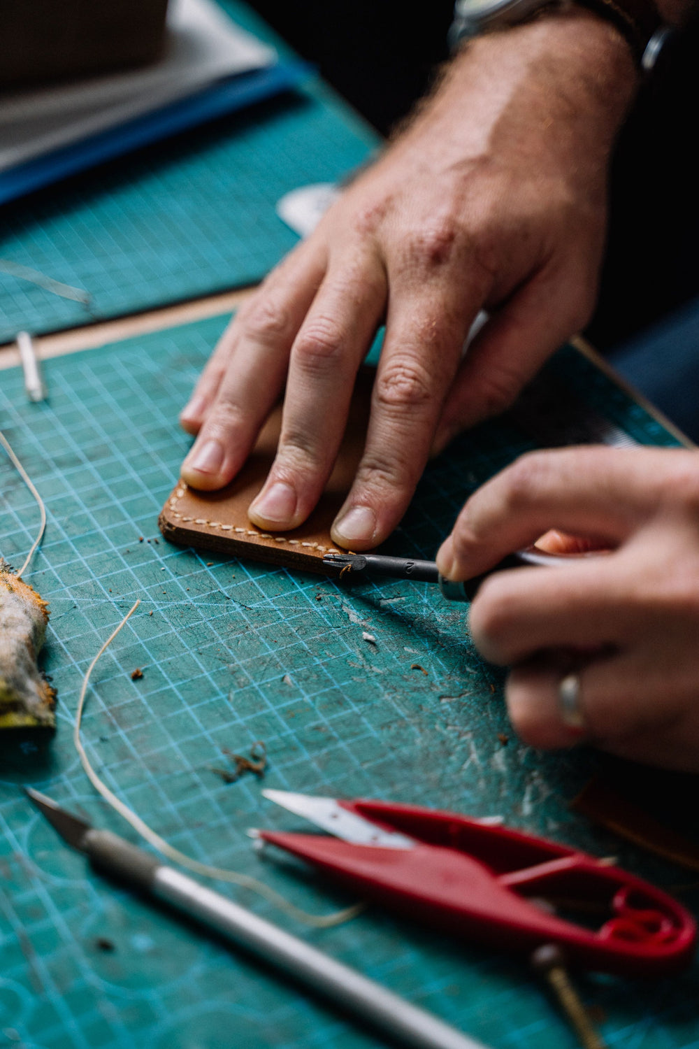 A leatherworker's hands stitching a hand-cut leather panel on a cutting mat, with awl, thread and snips laid out