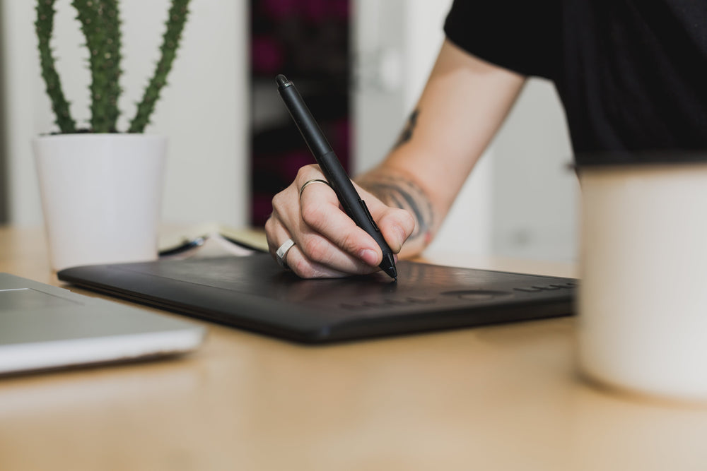 A designer drawing on a graphics tablet with a stylus, laptop and notebook alongside on the desk